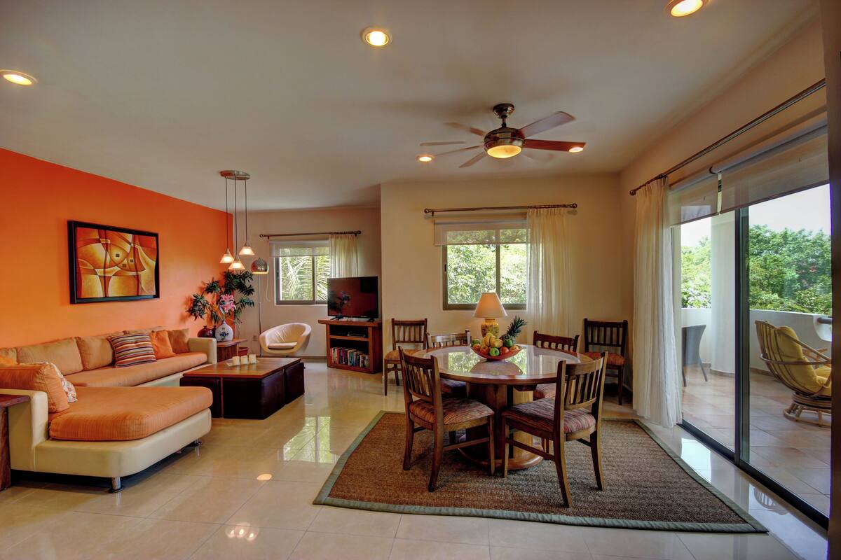 A spacious living room with a sofa, dining table, and chairs, a ceiling fan, and large windows with curtains.