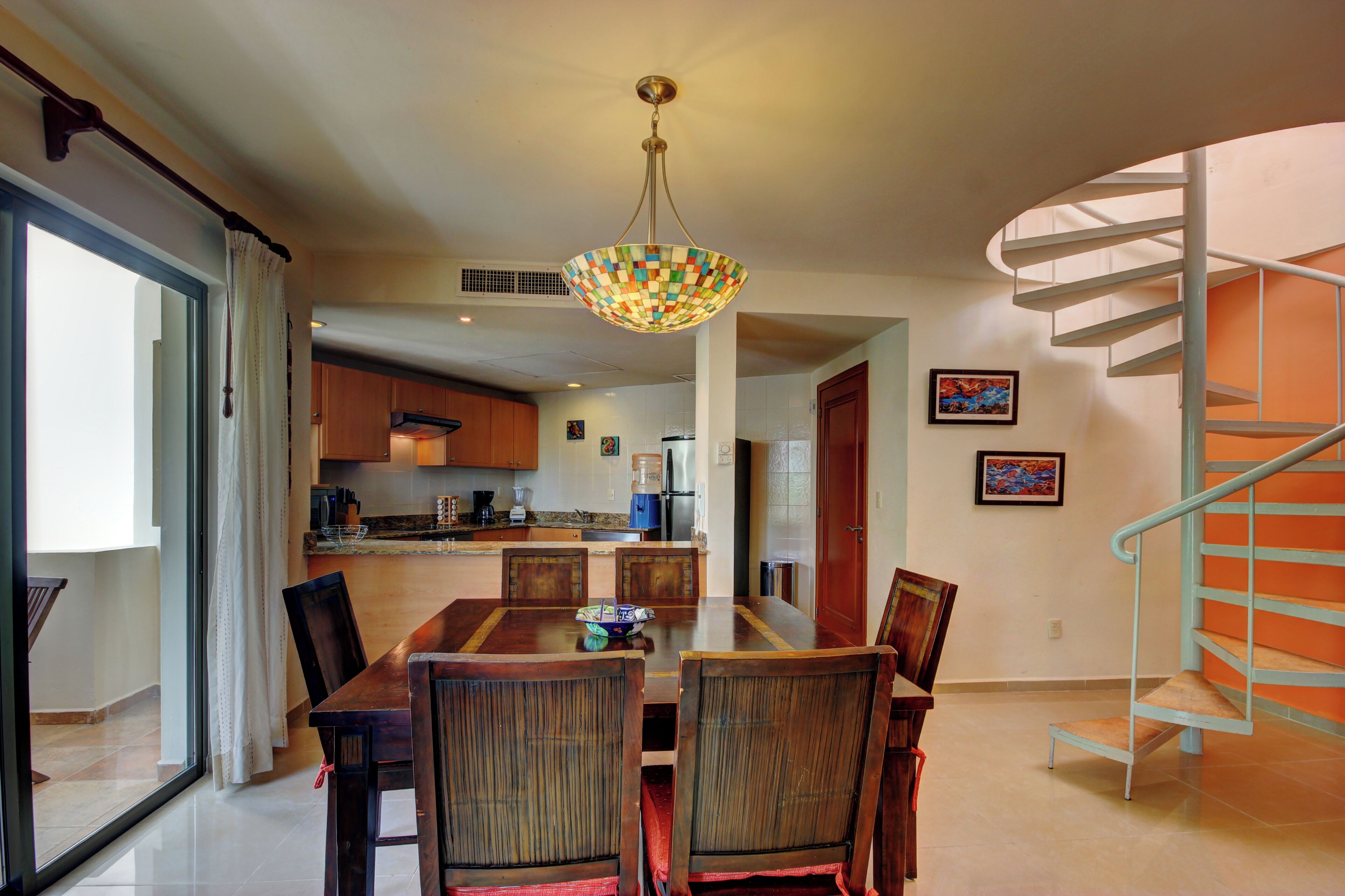 A dining area with a table and chairs, a kitchen in the background, and a spiral staircase.