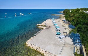 Eine Vogelperspektive zeigt einen Strand mit klarem, blauem Wasser, einem Betonsteg und Reihen von Liegestühlen mit Sonnenschirmen.