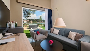 Camera d'albergo moderna con divano, tavolino, scrivania con lampada e vista su piscina e vegetazione.