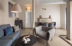 Ein modernes Wohnzimmer mit einem grauen Sofa, einem runden Couchtisch mit einer Auswahl an Gebäck und einem grauen Sessel. Es gibt eine Essecke mit Tisch und Stühlen, einen Kamin aus Stein und eine Tür zu einem weiteren Raum.