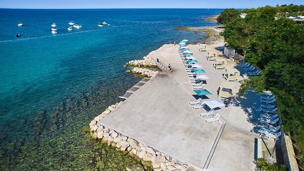 Vue aérienne d’une plage avec une eau d’un bleu limpide, une promenade en béton et des rangées de chaises longues sous des parasols.