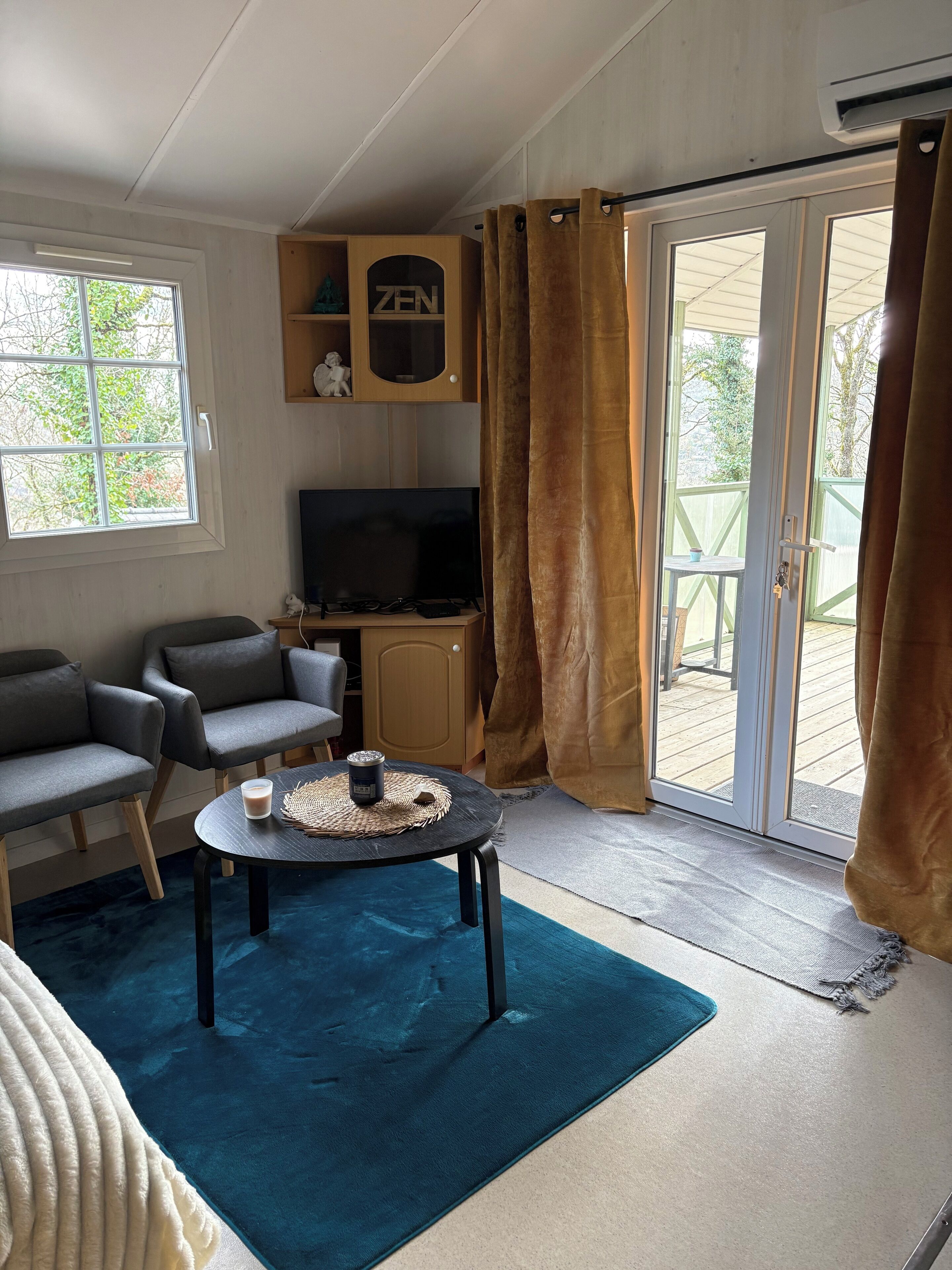 Ein Wohnzimmer mit Fernseher, zwei grauen Sofas, einem Couchtisch mit Kerze und einer Schiebetür zum Außenbereich.