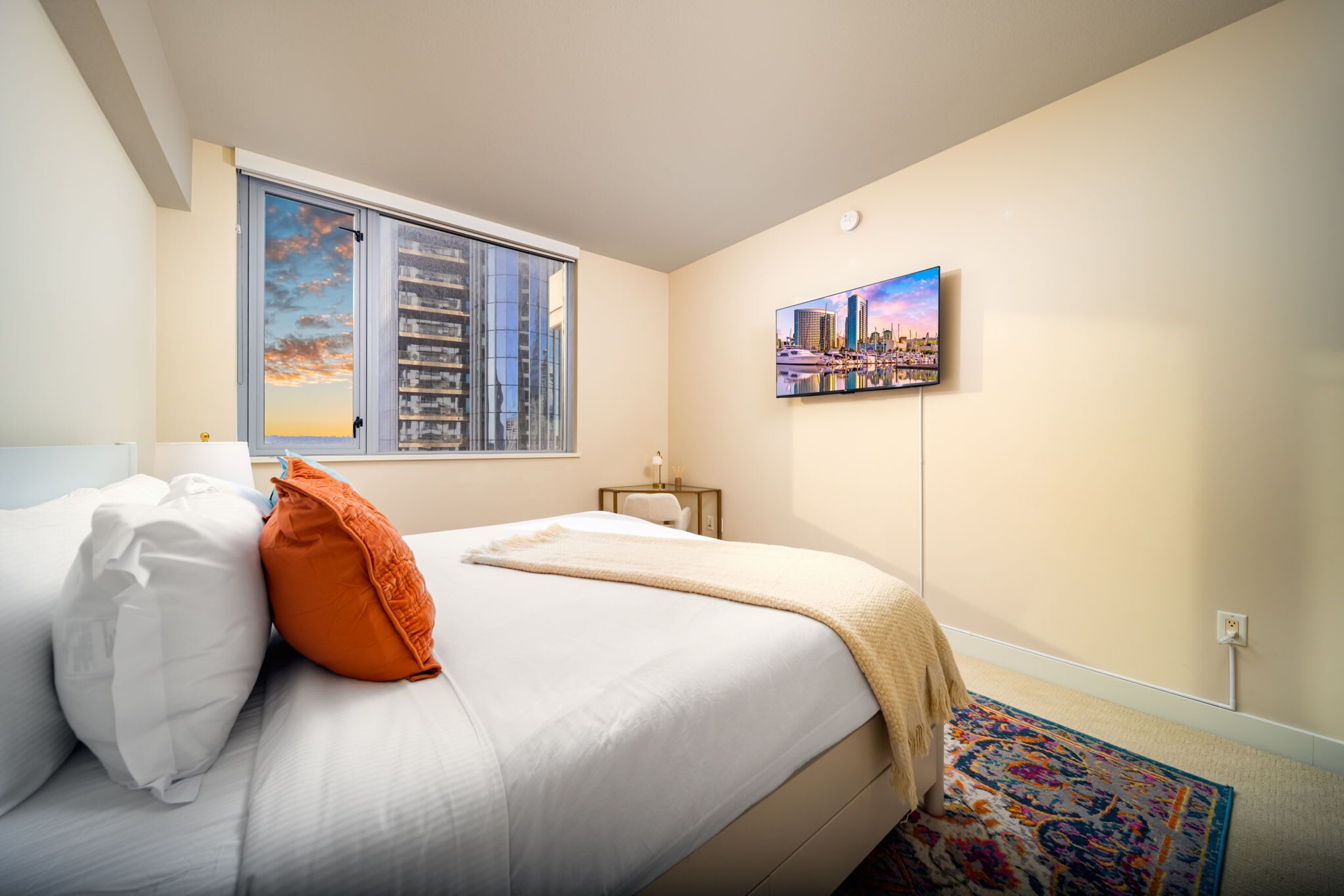 Une chambre à coucher avec un lit, des oreillers, une fenêtre donnant sur le paysage urbain, un tableau accroché au mur et une petite table avec une lampe.