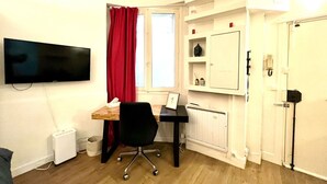Ein kleiner, gut beleuchteter Raum mit Schreibtisch, Stuhl und einem an der Wand befestigten Flachbildfernseher. Es gibt ein Fenster mit roten Vorhängen, eine Heizung und eine Tür mit Griff.