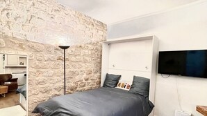 Ein Schlafzimmer mit Steinwand, einem Bett mit grauer Bettwäsche, einem an der Wand befestigten Fernseher und einem kleinen Nachttisch.