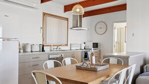 A modern kitchen with white cabinetry, a wooden dining table, and a hanging light fixture. - Serenity Sands - 34 Neighbour Ave, Goolwa Beach (Goolwa Beach)