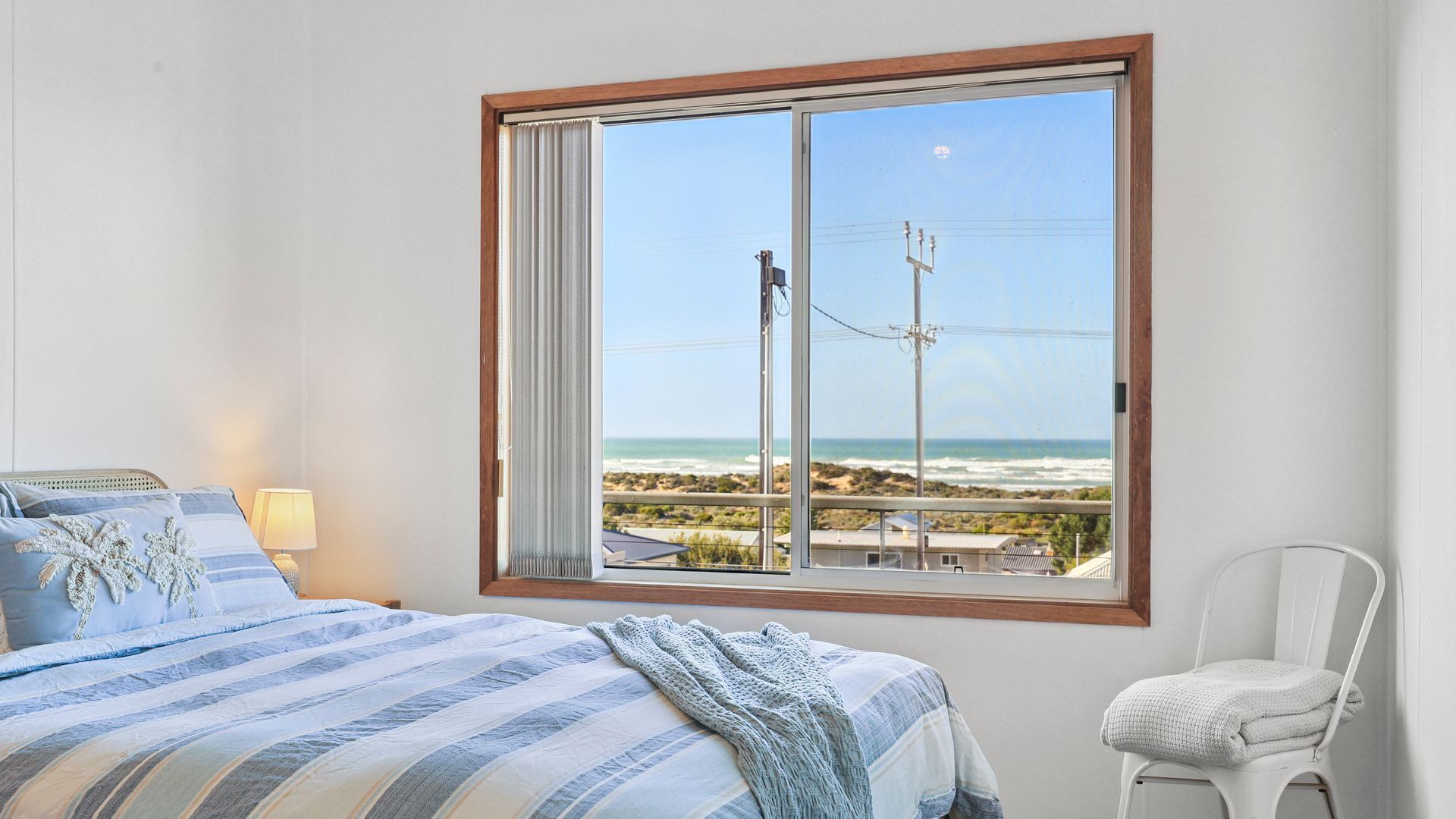 A bedroom with a large window overlooking a beach, a bed with striped bedding, and a chair.