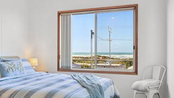 A bedroom with a large window overlooking a beach, a bed with striped bedding, and a chair.