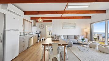 A modern kitchen with white appliances, a dining area with a wooden table and chairs, a living room with a sofa and a view of the outdoors.