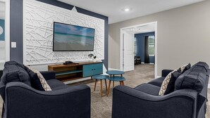 Un salon moderne avec un grand téléviseur, des canapés sombres, une table basse bleue et un panneau mural décoratif blanc.
