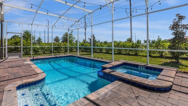 Une piscine couverte avec un jacuzzi, entourée d’une structure métallique et de panneaux de verre, aménagée dans un espace paysager agrémenté d’arbres et offrant une vue sur un ciel dégagé.
