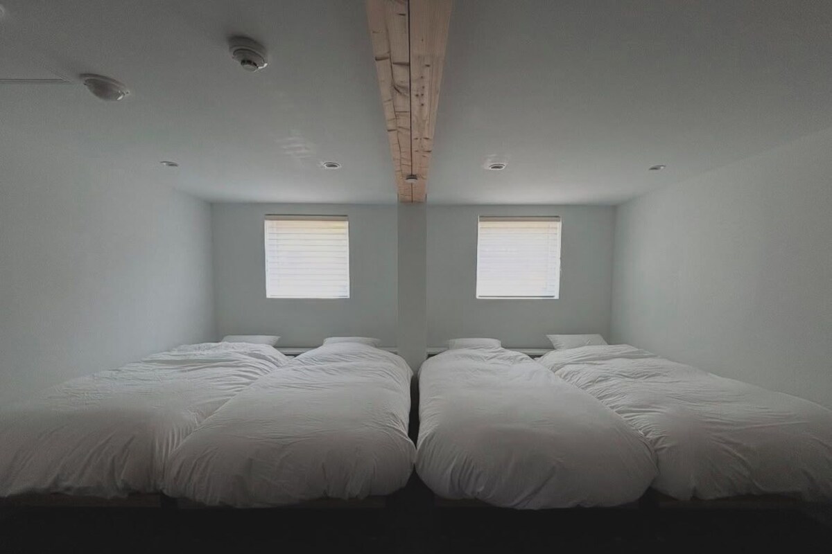 A room with two beds, a wooden ceiling beam, and two windows with blinds.