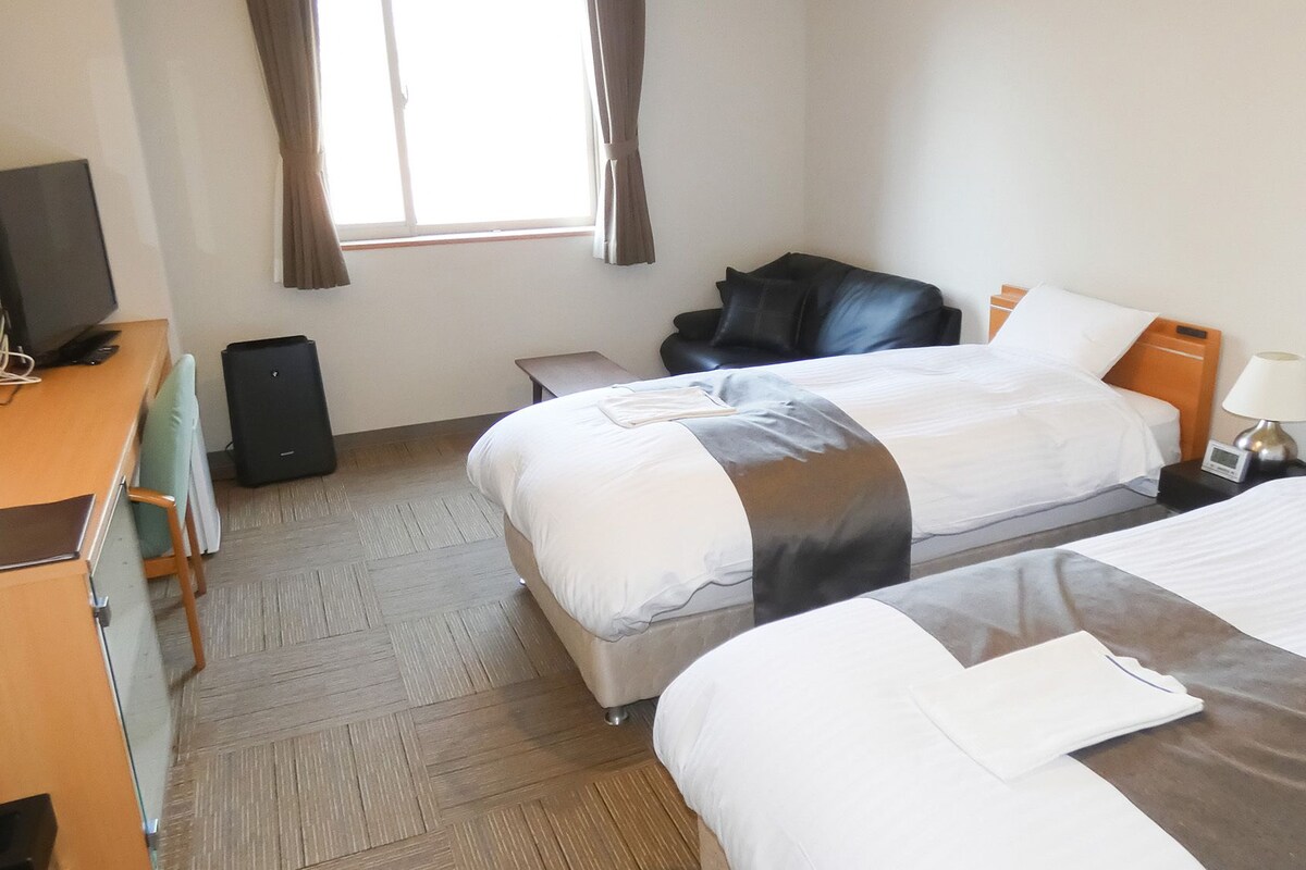 Une chambre d’hôtel avec deux lits, un bureau, une chaise, une télévision et une fenêtre avec des rideaux.