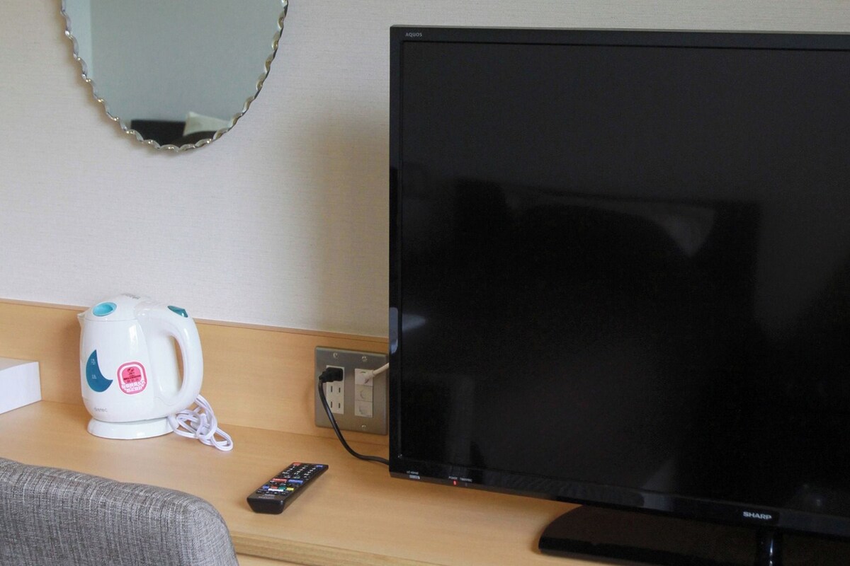 Een hotelkamer met een flatscreen televisie, een witte elektrische waterkoker en een afstandsbediening op een houten tafeltje.