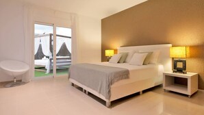 A modern bedroom with a large bed, bedside lamps, a white chair, and a view of a building through a glass door. - Villa Julieta – Ibiza Town (2 min) (Nuestra Señora De Jesus)