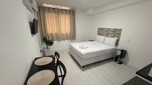 Ein kleines Zimmer mit einem Bett, einem kleinen Tisch mit zwei Stühlen, einer Wand-Klimaanlage und einem fensterbepflanzten Fenster.