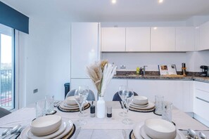 A modern dining area with a table set for four, white cabinets, and a kitchen in the background. - Stylish duplex 2-bed with balcony & parking (Manchester)