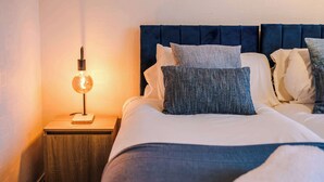 A neatly made bed with a dark headboard, white bedding, and several pillows. A small wooden nightstand with a lamp is visible to the left. - 5BR Contractor House | Free Parking | Near Airport (Manchester)