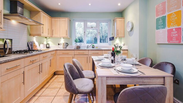 A modern kitchen with wooden cabinets, a dining table set for four, and a view of the outdoors through a window. - 5BR Contractor House | Free Parking | Near Airport (Manchester)