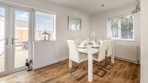 Une salle à manger avec une table blanche dressée pour quatre personnes, des chaises blanches, un radiateur blanc et une fenêtre avec des stores.