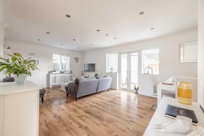 A spacious living area with a grey sofa, wooden flooring, a kitchen area, and large windows. - Large 6 Bed Maidenhead Stay With Free Parking (Maidenhead)