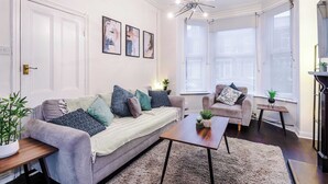 Ein Wohnzimmer mit einer Couch, einem Couchtisch und zwei Beistelltischen. An der Wand hängen drei gerahmte Bilder und eine Kristallleuchter.