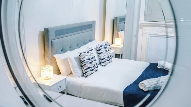 Ein ordentlich gemachtes Bett mit grauem Kopfteil, zwei gemusterten Kissen, einem weißen Nachttisch mit Lampe und einer blauen Decke.