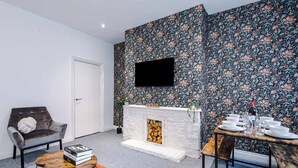 Uma sala de estar moderna com lareira, TV de tela plana na parede, mesa de jantar para quatro pessoas e um espaço de estar acolhedor com sofá e pequena mesa de centro.