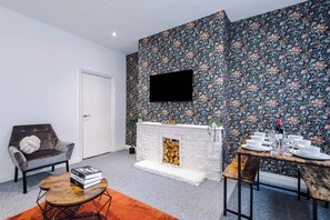 A modern living room with a fireplace, a flat-screen TV mounted on the wall, a dining table set for four, and a cozy seating area with a sofa and a small coffee table. - 3 bedroom town house - Etihad Stadium - Free Parking (Manchester)