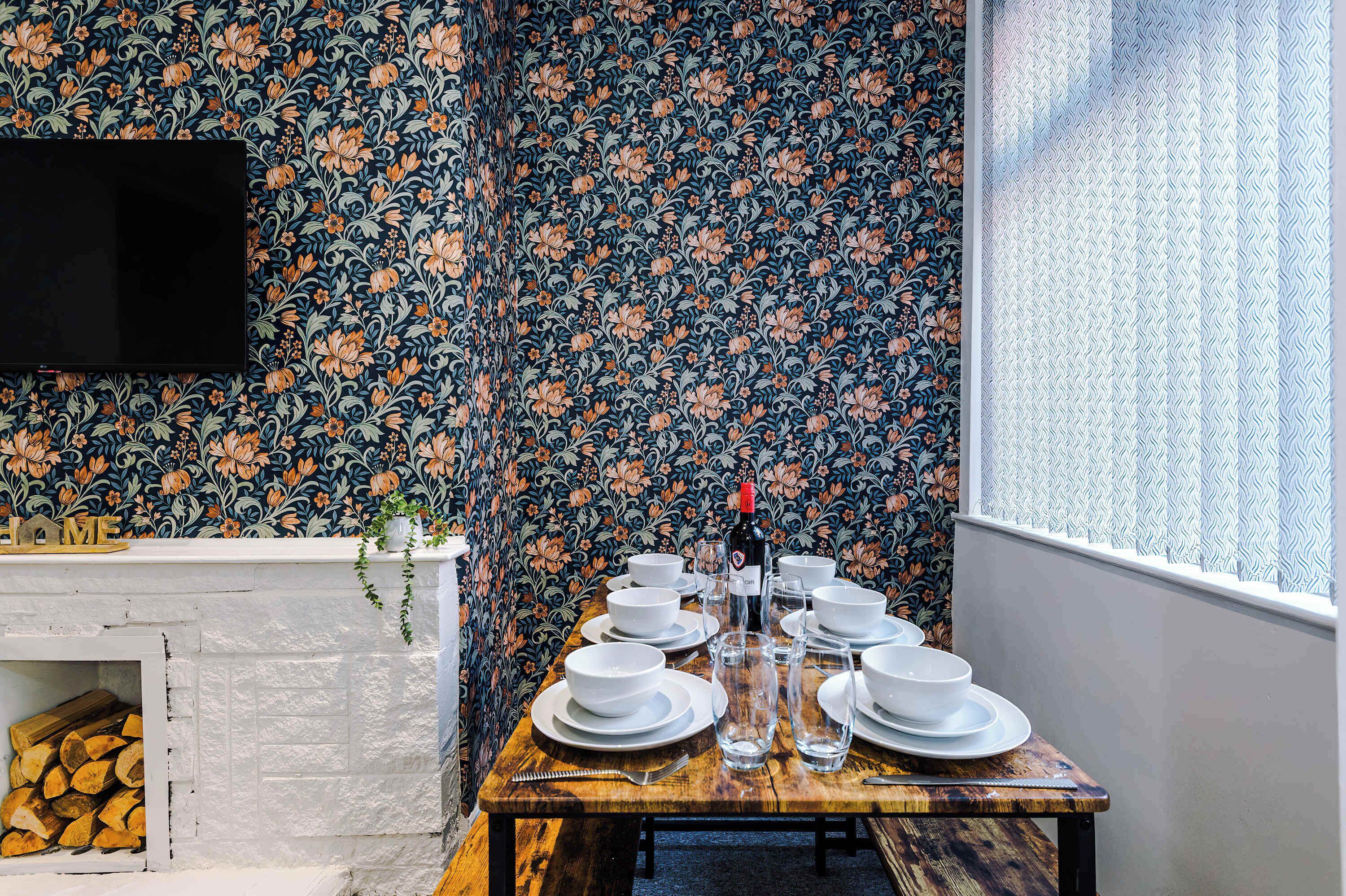 A dining table set with white plates and bowls, a bottle, and glasses, against a floral wallpaper backdrop.