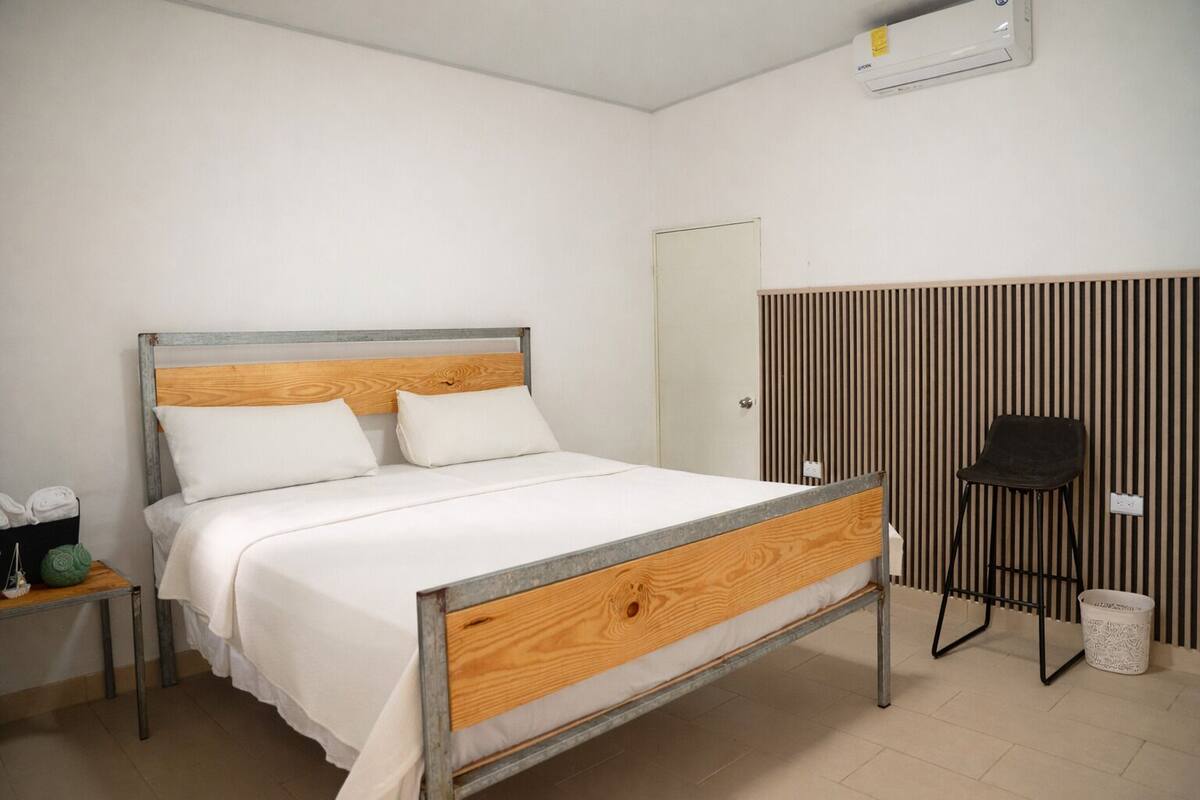 Uma cama de solteiro com cabeceira de madeira e estrutura metálica, uma mesinha de cabeceira com um cesto, uma cadeira e um ar-condicionado fixado na parede.