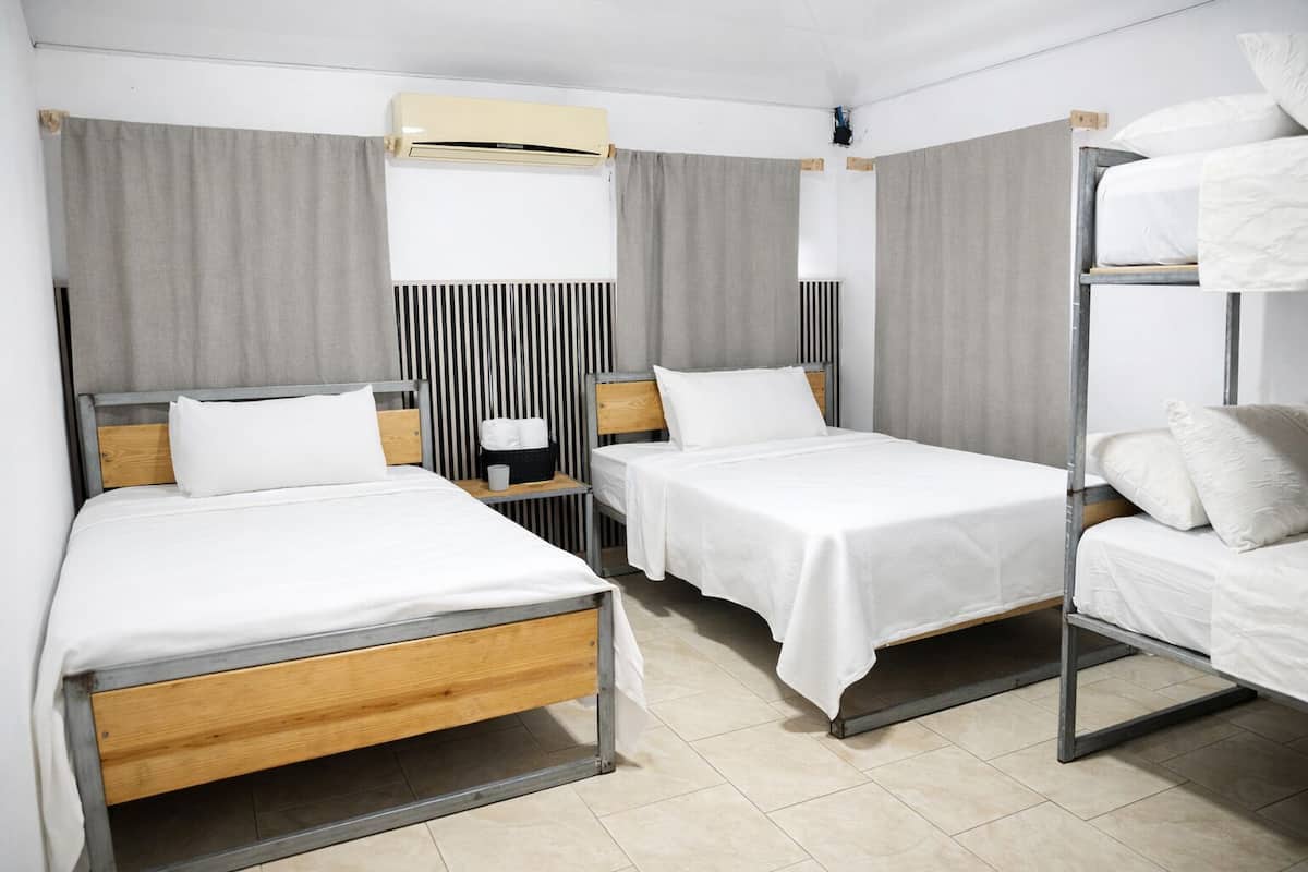 Quarto de hotel com duas camas de solteiro, uma cama beliche, uma mesinha de cabeceira com abajur, ar condicionado fixado na parede e um painel decorativo listrado.