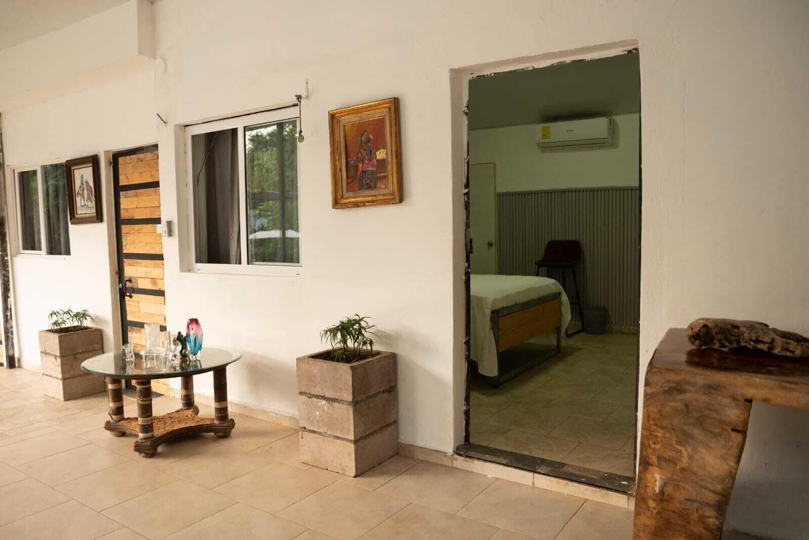 Una habitación con una cama, una mesita con objetos decorativos, una ventana y una puerta que da a otra habitación.