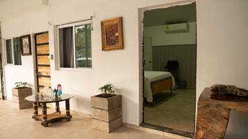 Una habitación con una cama, una mesita con objetos decorativos, una ventana y una puerta que da a otra habitación.