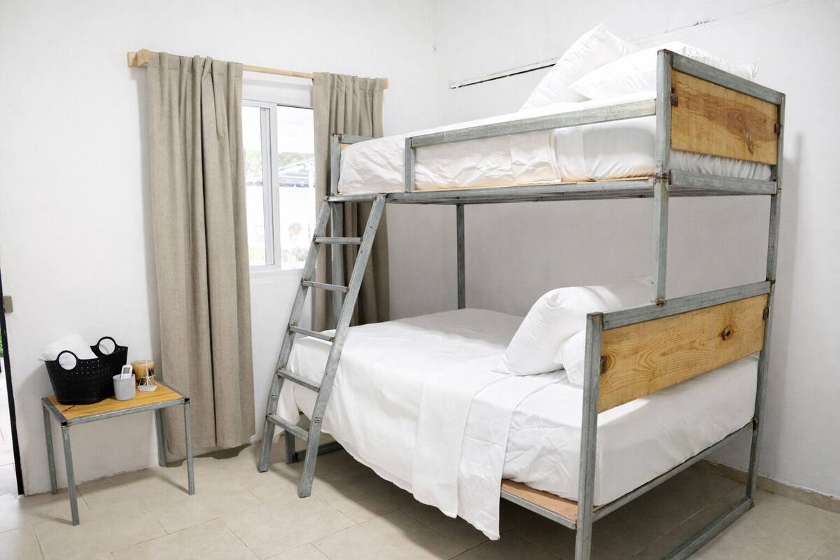 Uma cama beliche com escada, uma mesinha com cesto e vela, e uma janela com cortinas.