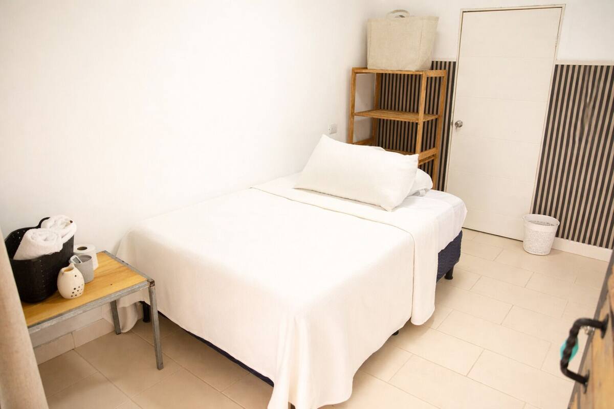Uma cama de solteiro com roupa de cama branca, uma pequena mesa de cabeceira com luminária, uma estante listrada em preto e branco e uma lixeira branca.