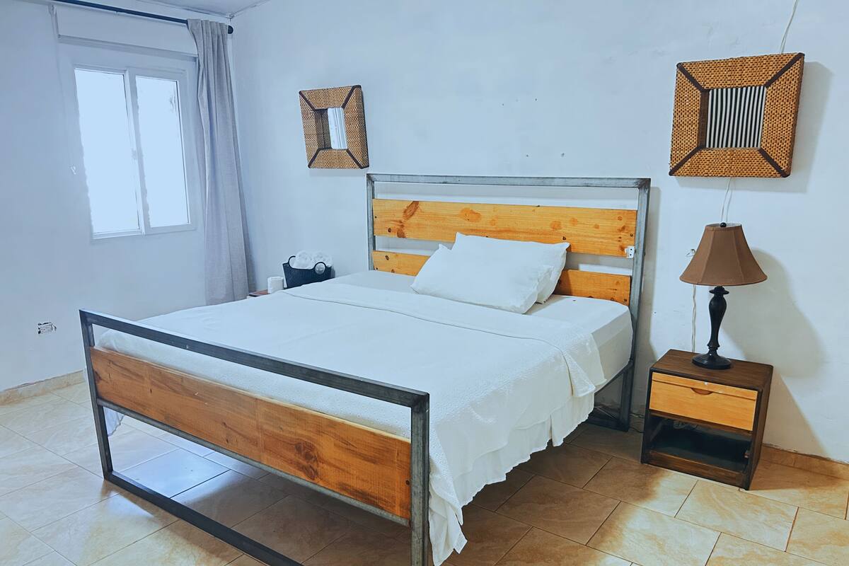 Quarto com estrutura de cama de madeira, roupa de cama branca, mesa de cabeceira com abajur e dois objetos decorativos fixados na parede.