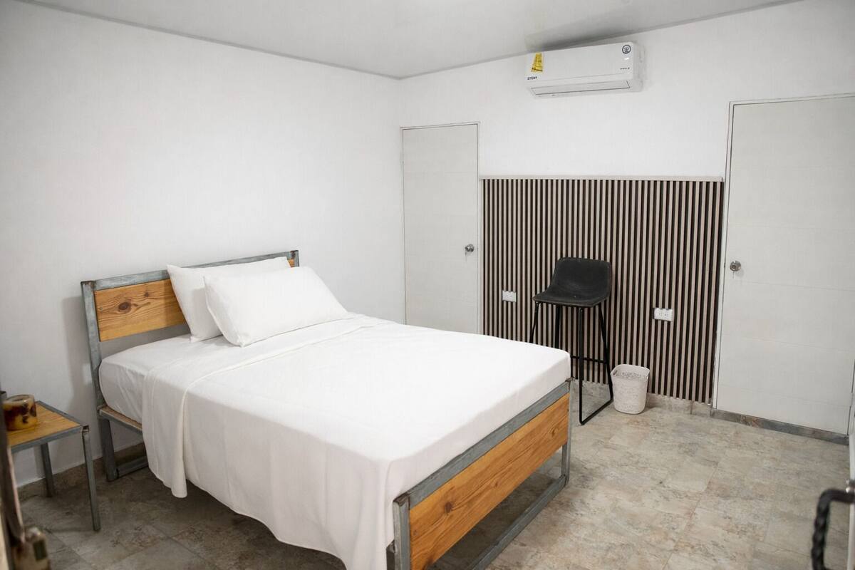 Um quarto com cama, uma cadeira, uma mesinha, um radiador e uma porta.