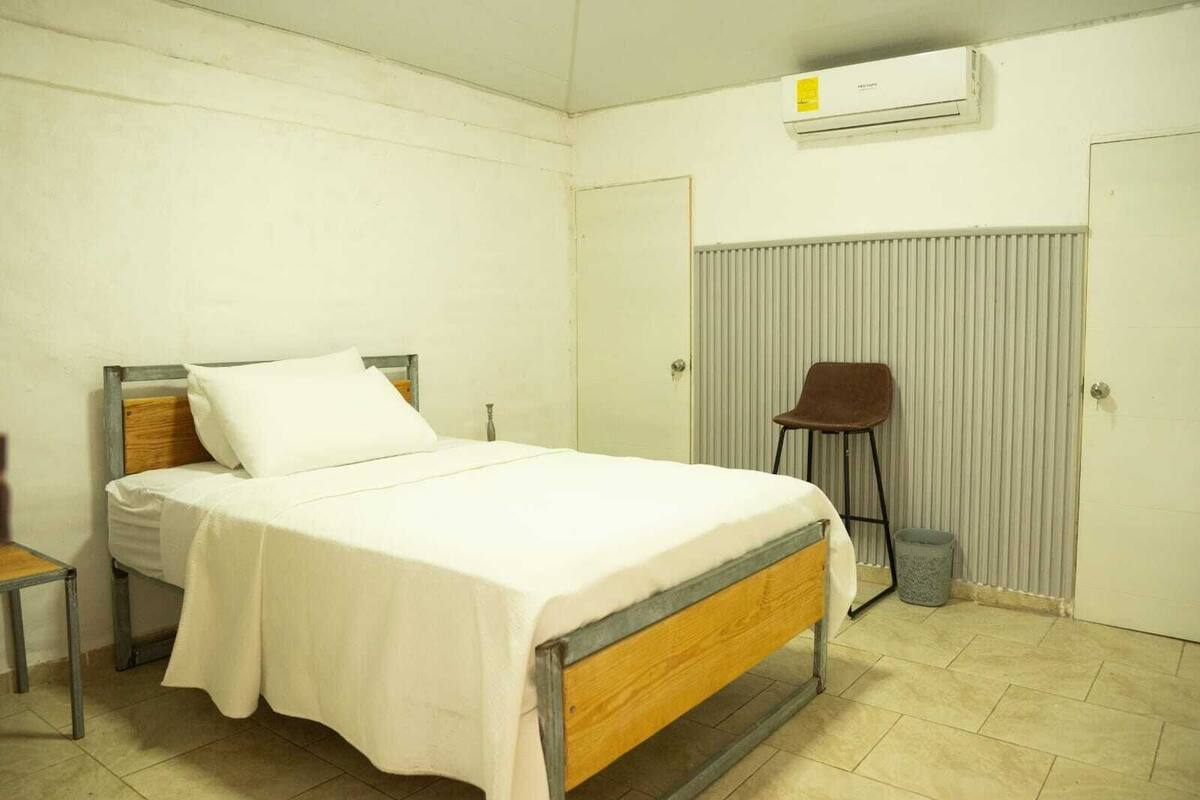 Quarto individual com cama de solteiro, cabeceira de madeira, colcha branca, cadeira marrom, radiador e ar condicionado.