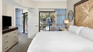 A hotel room with a bed, a TV, a desk with a lamp, and a view of an outdoor pool area. - Experience Family Fun and Coastal Charm at Marriott's Barony Beach- 2 Bd Garden (Hilton Head Island)
