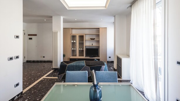 Modernilla designilla sisustettu hotellihuone, jossa on lasinen ruokapöytä, sohvaryhmä, seinään upotettu televisio ja kattovalaisin.
