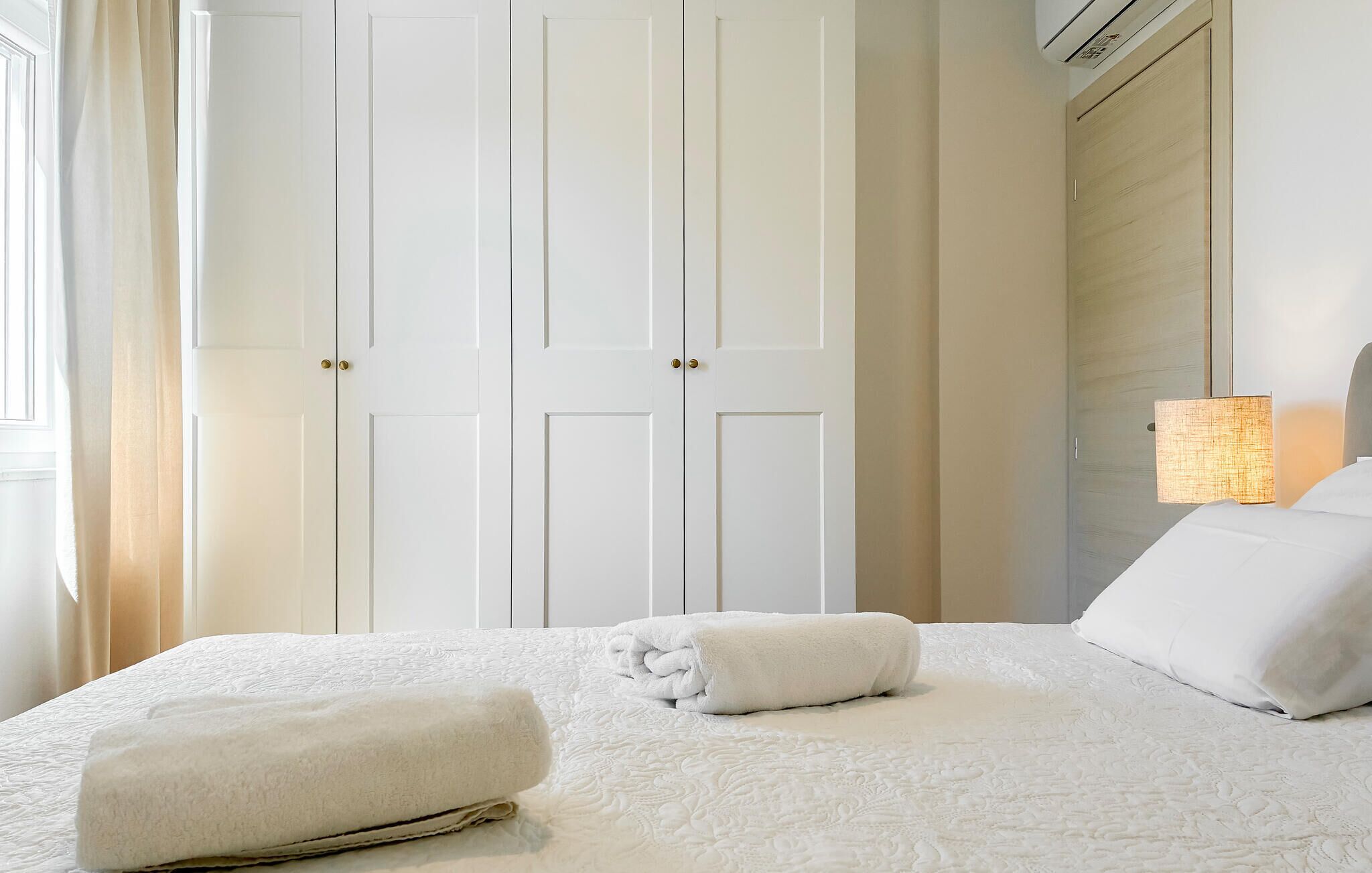 A hotel room with a bed, two white towels, a nightstand, a lamp, and a wardrobe.
