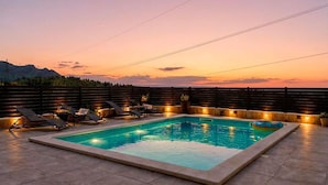 Een rechthoekig zwembad met ligbedden en een jacuzzi, omgeven door een houten omheining en met uitzicht op de bergen bij zonsondergang.