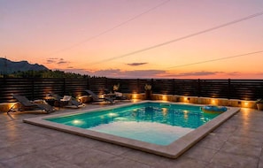 A rectangular swimming pool with lounge chairs and a hot tub, surrounded by a wooden fence and a view of the mountains at sunset. - Beautiful home in Klis (Klis)