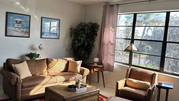A living room with a sofa, armchair, ottoman, and a large window with a view of palm trees. - Gorgeous 1-bedroom condo close to work and play only steps to beach (Satellite Beach)