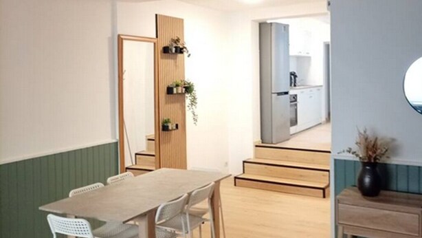 Un coin repas avec une table en bois et des chaises blanches, une porte en bois dotée d’étagères, et une vue sur un espace cuisine.