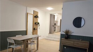 Un coin repas avec une table et des chaises en bois, une petite table d’appoint avec un vase, et une cuisine en arrière-plan.