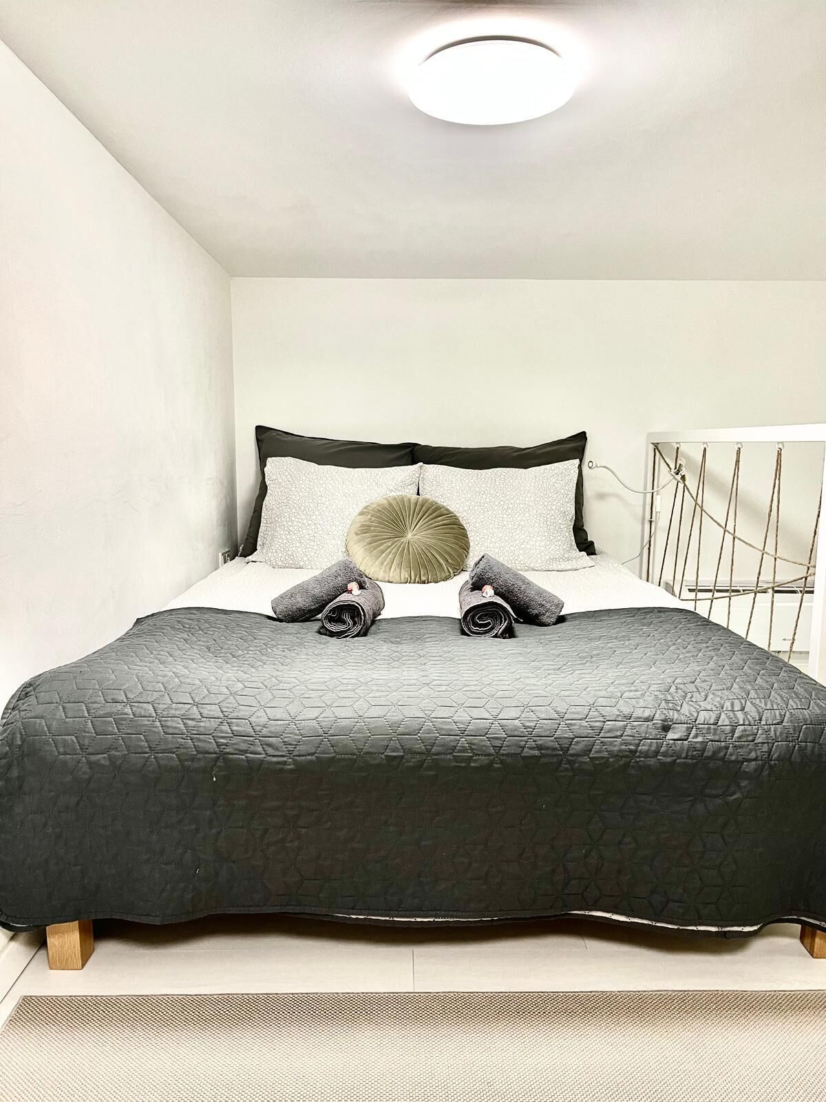 Un lit bien fait, avec une couverture sombre, deux serviettes enroulées et un coussin décoratif.
