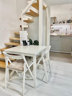 A white dining table with two chairs, a wooden staircase, and a kitchen area with white cabinets and a tiled backsplash. - Central BUDAPEST Studio ⭐️| AC+Netflix (Budapest)